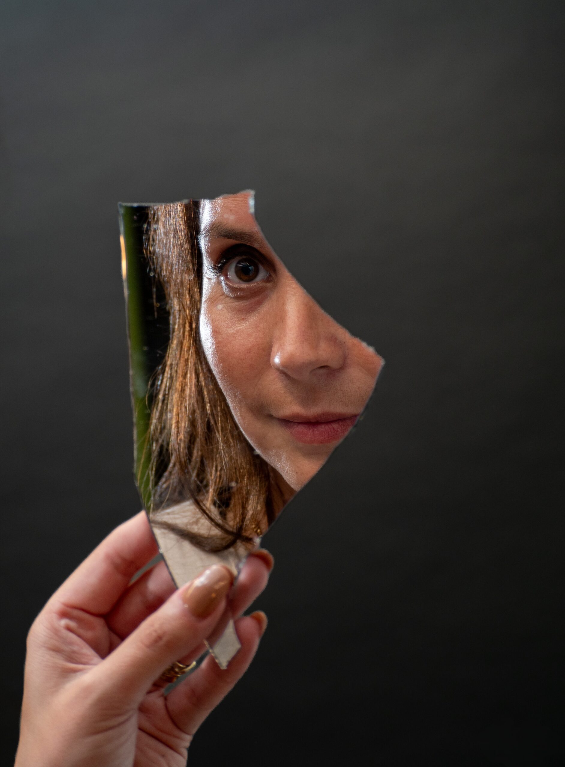 Vrouw houdt gebroken spiegel vast – haar oog weerspiegelt het thema van zelfreflectie en identiteit.