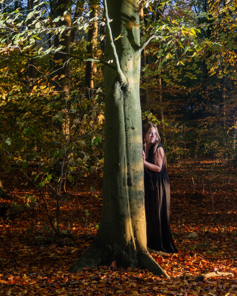 Fotografie van een meisje verscholen achter een boom in het bos, waardoor een mysterieuze en dromerige sfeer ontstaat. Deze foto legt de verbondenheid van het meisje met de natuur vast in een moment van verborgen aanwezigheid.